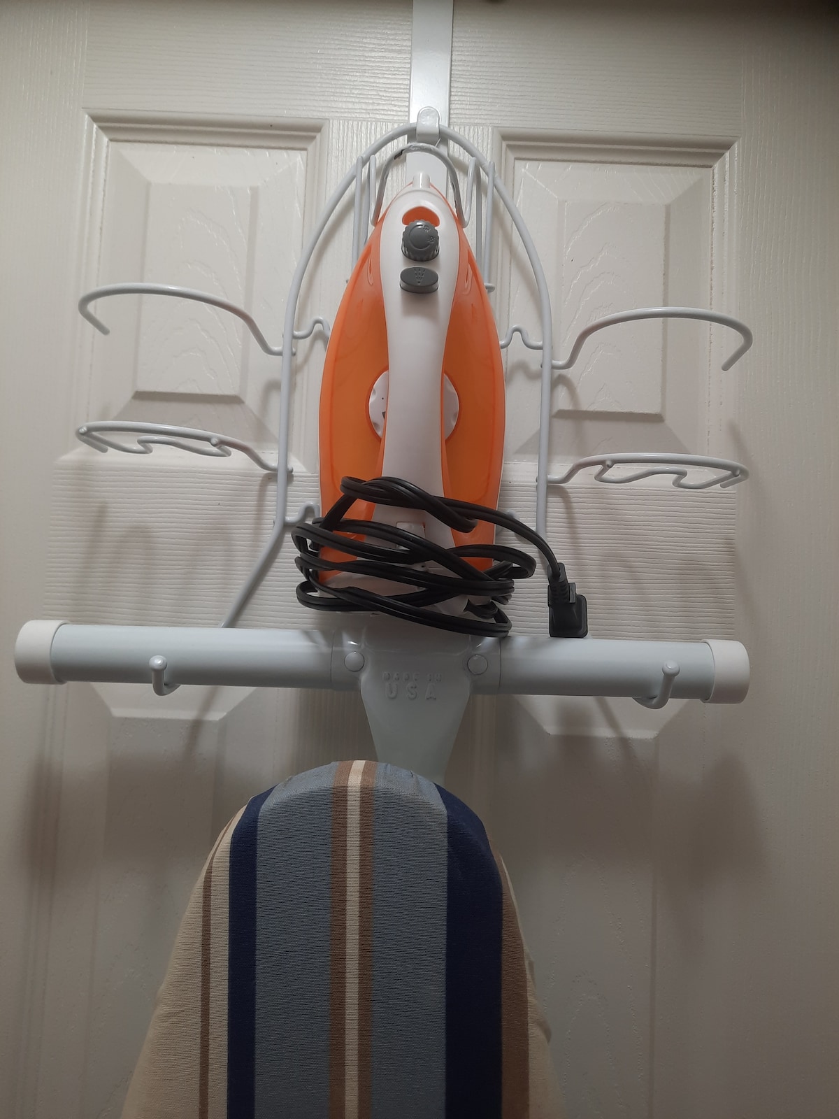 An orange steam iron is mounted on a wall-mounted holder, with its cord neatly wrapped. Below, an ironing board with a striped cover is visible, providing a practical element for guests' convenience.