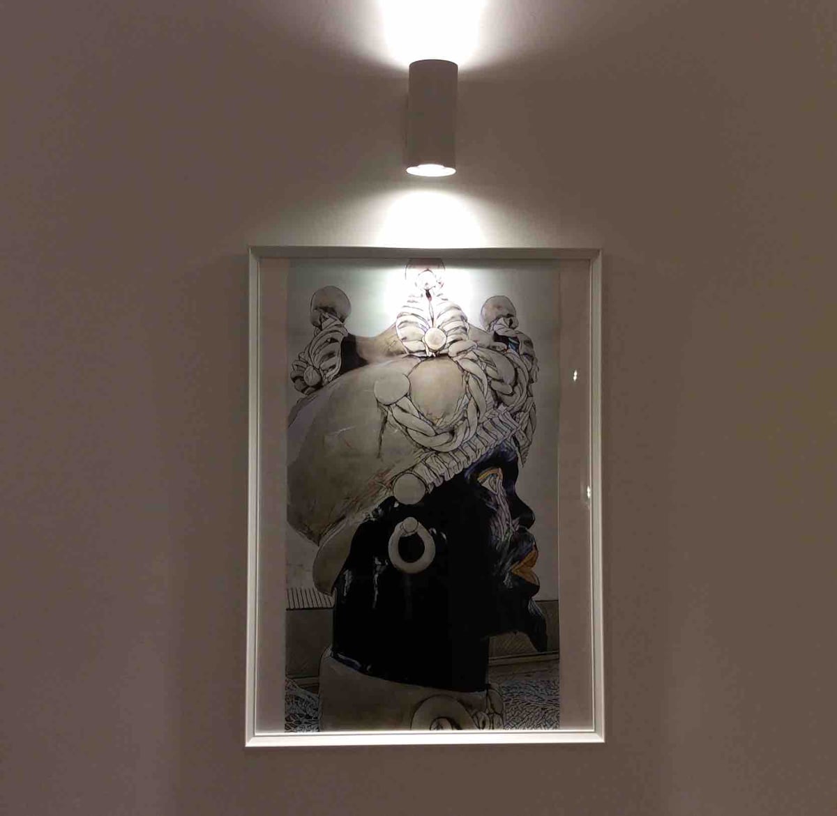 A framed artwork is displayed on a light-colored wall, illuminated by a ceiling-mounted light fixture. The image features a portrait of a figure adorned with elaborate headgear, showcasing intricate details and textures that can be appreciated up close.