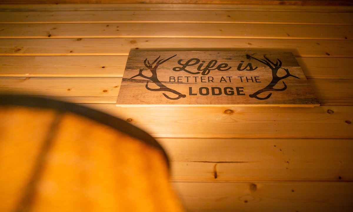 A wooden sign is displayed on a wall, featuring the phrase 'Life is better at the Lodge' surrounded by antler graphics. The warm light from a nearby lamp casts a gentle glow, illuminating the natural wood paneling of the interior.