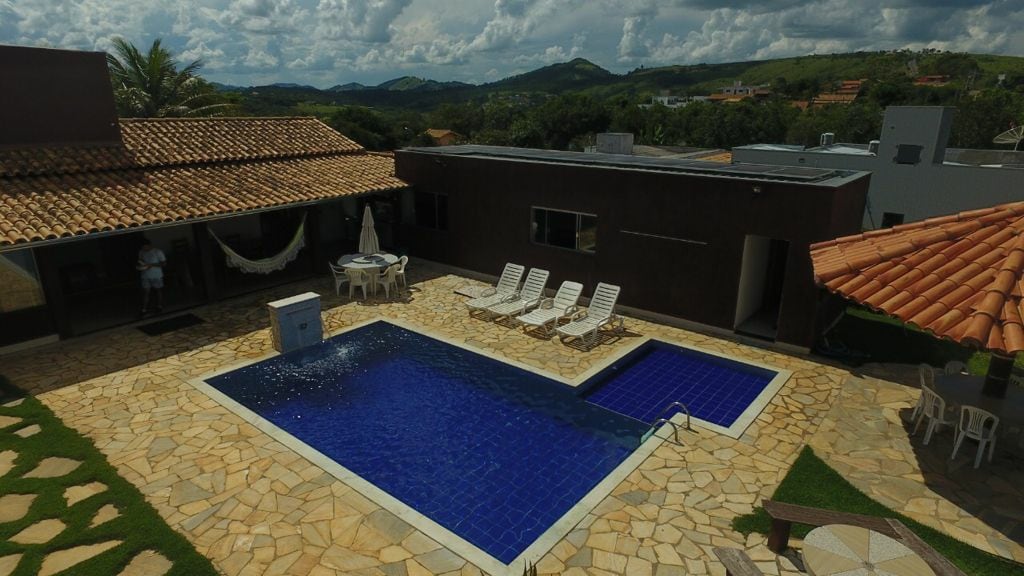 An outdoor area features a large swimming pool with a rich blue surface and a shallow section. Surrounding the pool are several lounge chairs positioned on a stone patio. A shaded pergola provides seating, and picturesque hills are visible in the background.