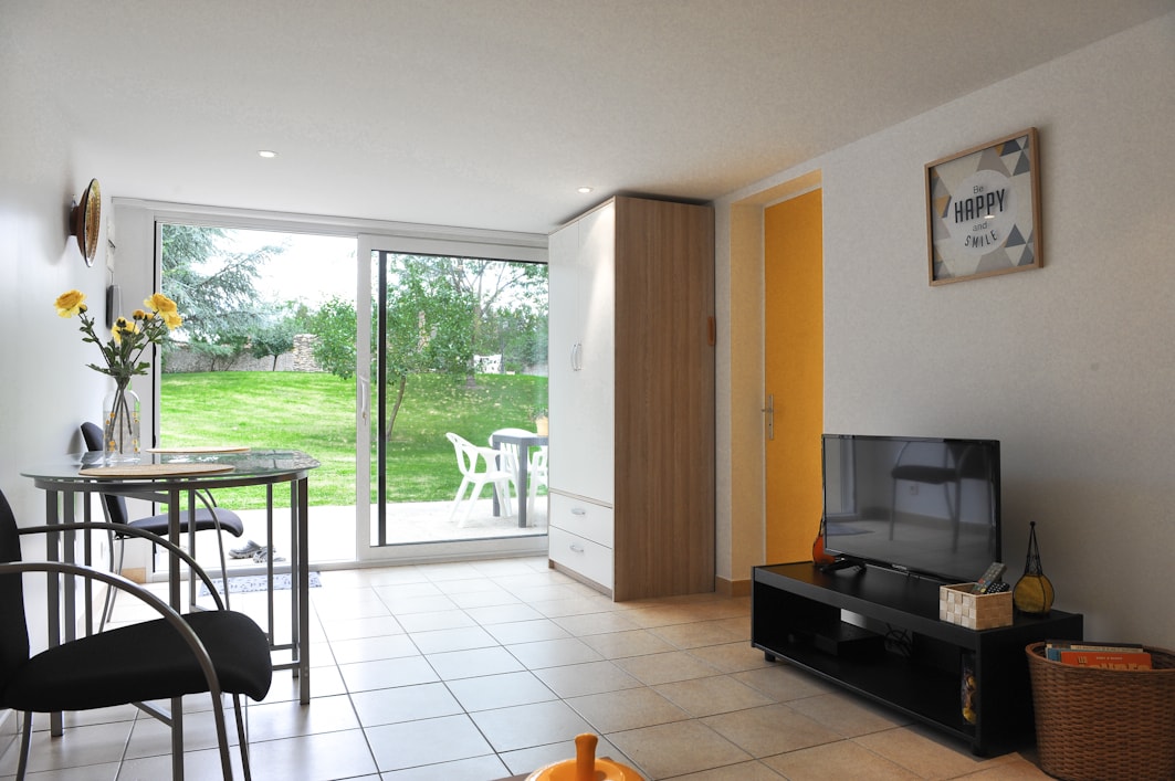 A bright living space features a glass sliding door that opens to a grassy outdoor area. A small dining table with two chairs is placed beside a television unit. Natural light illuminates the room, enhancing the clean, simple decor.