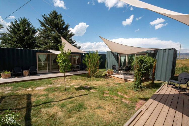 The outdoor area features two rustic shipping containers with large windows, shaded by fabric awnings. Comfortable seating is arranged on the deck, surrounded by greenery and a grassy space. The distant mountains and blue sky create a serene backdrop.