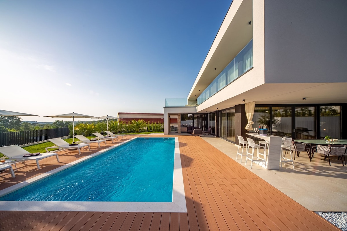 An outdoor view of the villa features a turquoise swimming pool surrounded by sun loungers and umbrellas. The modern architecture of the villa is highlighted with large glass doors leading from the interior to the patio area, creating an inviting atmosphere for relaxation and leisure.