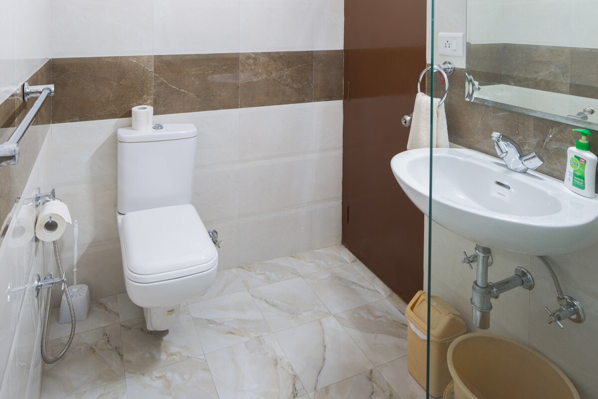 An en-suite bathroom is shown, featuring modern fixtures including a toilet and sink. Light-colored tiles cover the walls and floors, complemented by a contrasting brown accent strip. A glass shower enclosure is visible, along with essential toiletries and a roll of toilet paper neatly placed.