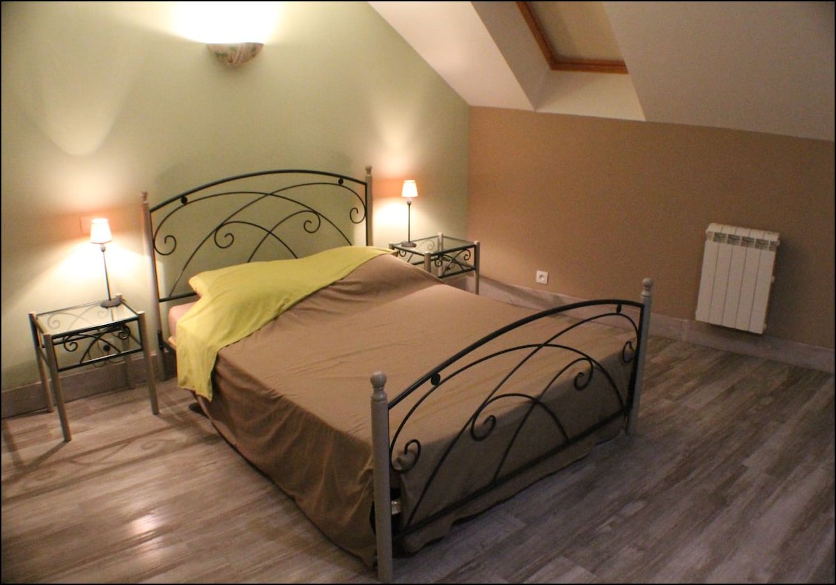 A spacious sleeping area features a metal-framed bed with neutral bedding and vibrant yellow accents. Nightstands with soft lighting are positioned on either side. Wooden flooring enhances the room's warmth, while sloped ceilings add a cozy character.