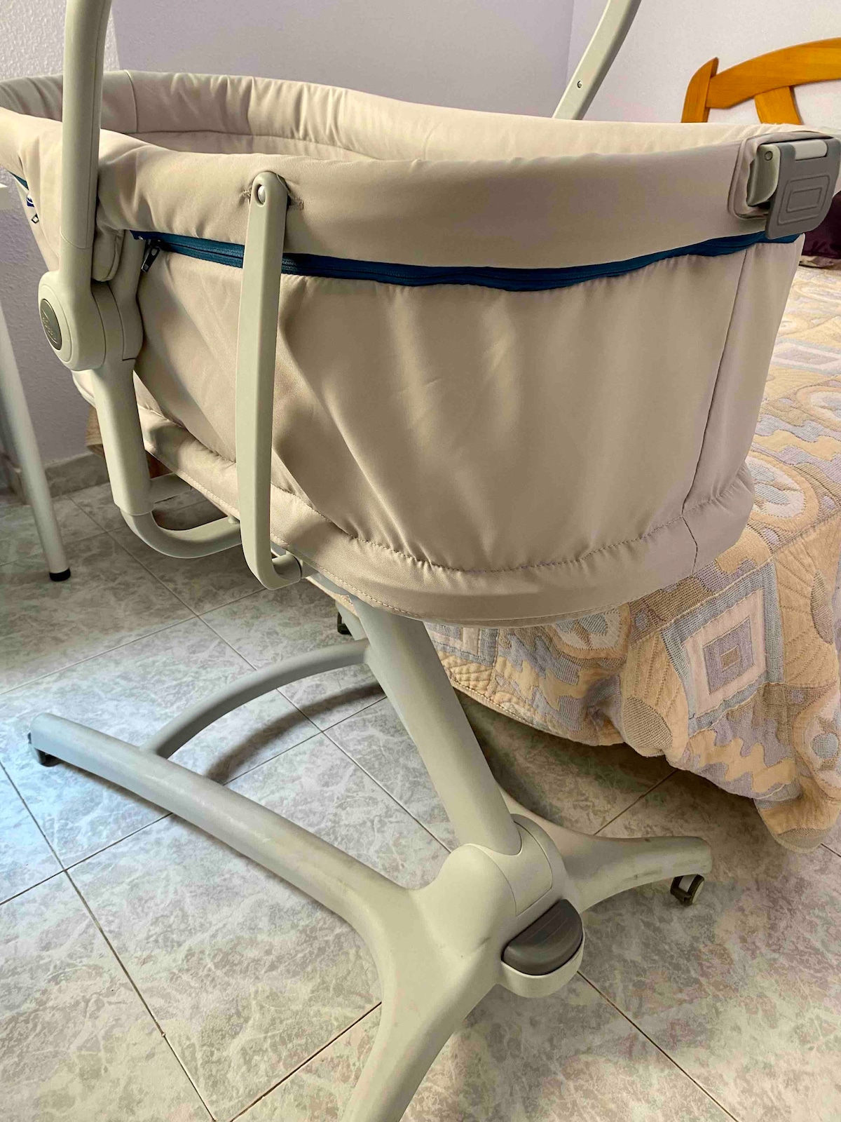 A bedside crib is positioned next to a spacious bed. The crib features a soft fabric interior and is mounted on a wheeled base for easy mobility. The bedding on the bed showcases a light pattern, harmonizing with the neutral tones of the crib.