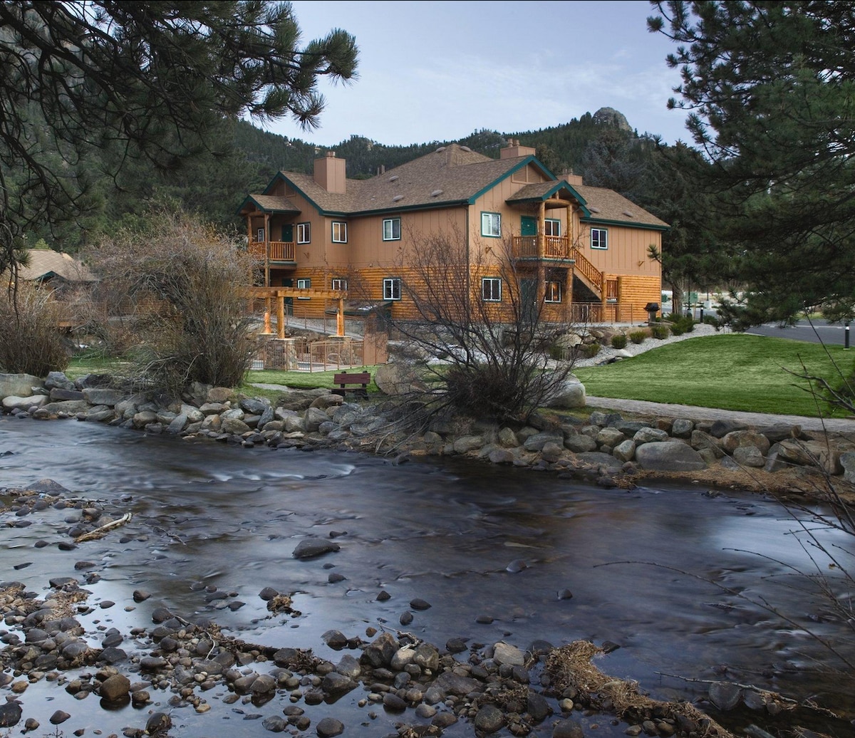 The image captures a scenic view of the building adjacent to a gently flowing river. Rocky hills and trees surround the area, providing a natural backdrop. Details such as wooden balconies and stone pathways are visible, illustrating the property's integration with its outdoor setting.