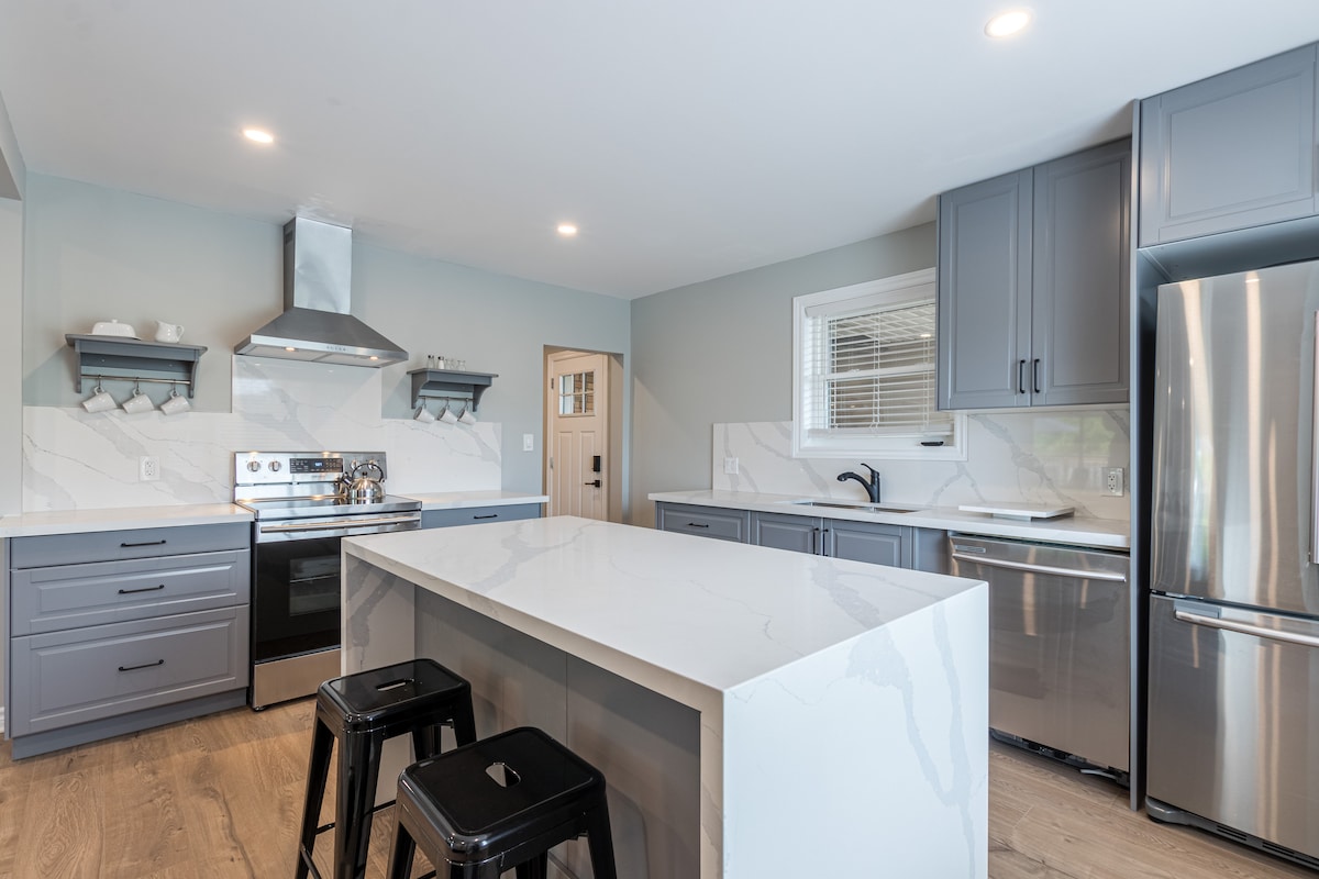 The modern kitchen features sleek gray cabinets and a large center island with a white quartz countertop. Stainless steel appliances, including an oven and refrigerator, complement the space. Bright lighting enhances the clean lines and ample work surfaces, creating a functional area for meal preparation.