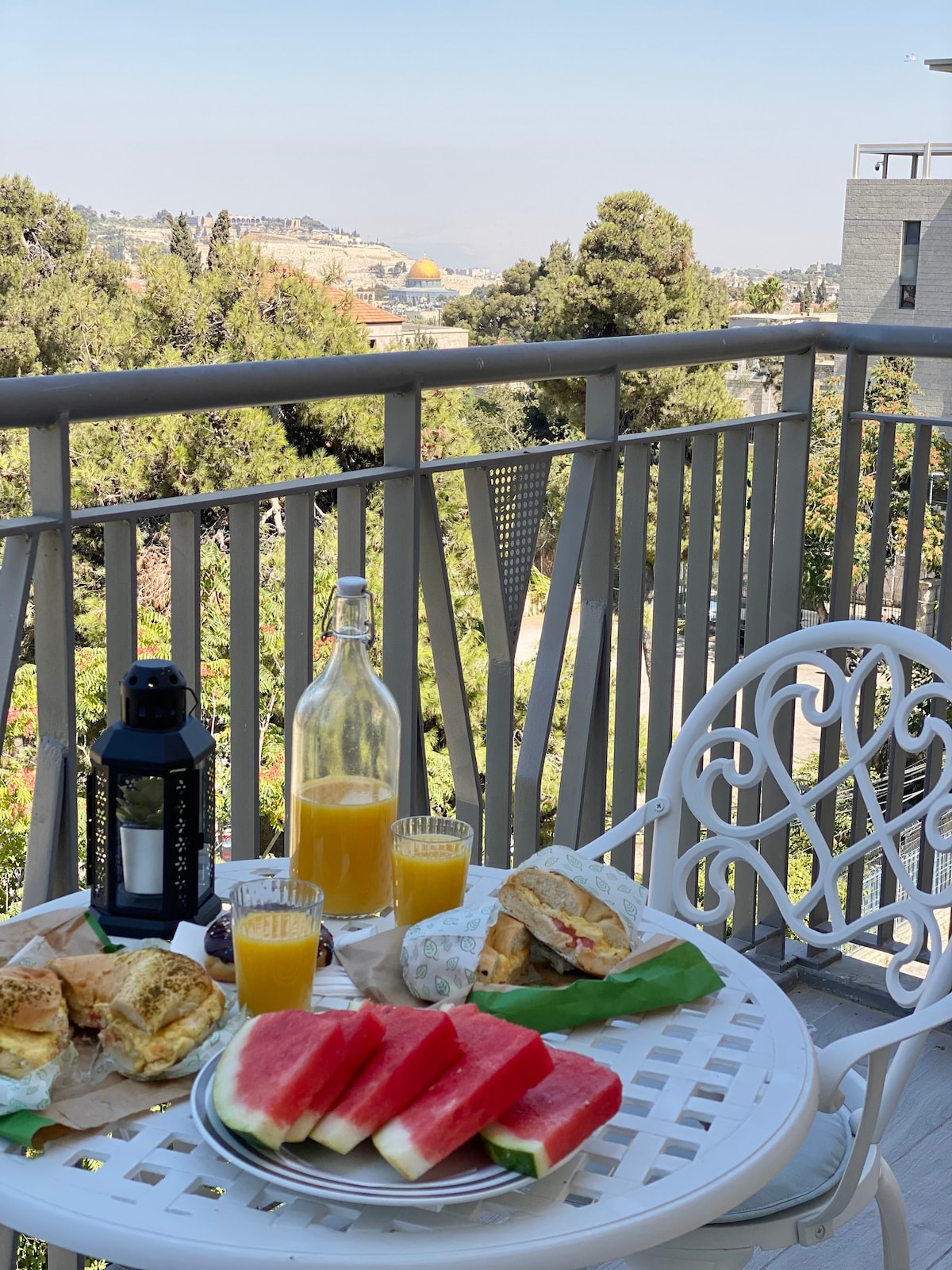 The balcony features a round table set with a plate of sliced watermelon, freshly made sandwiches, and two glasses of orange juice. A lantern and a bottle add to the inviting atmosphere, surrounded by views of greenery and the distant Temple Mount.