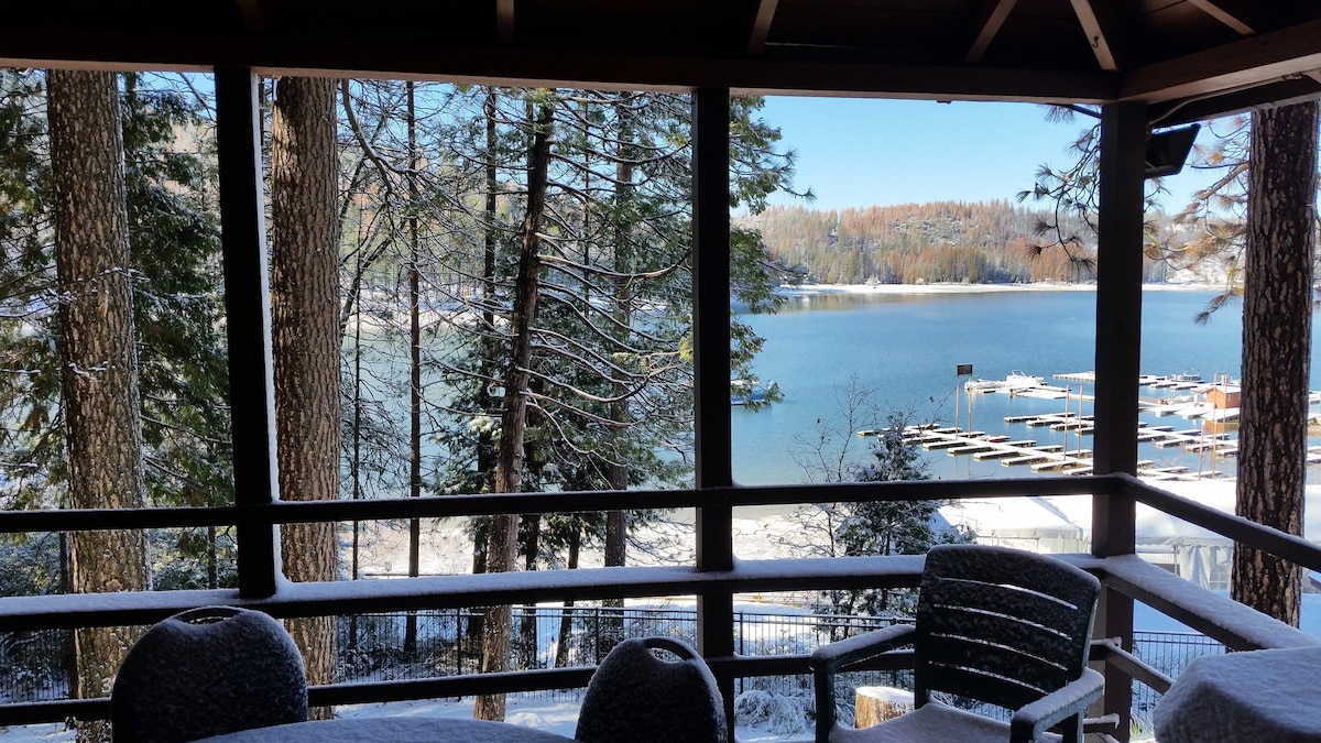 A covered outdoor patio provides a view of Bass Lake through large windows. Trees frame the scene, with the calm water reflecting a clear sky. A dining table and chairs are visible on the patio, offering seating to enjoy the surroundings.