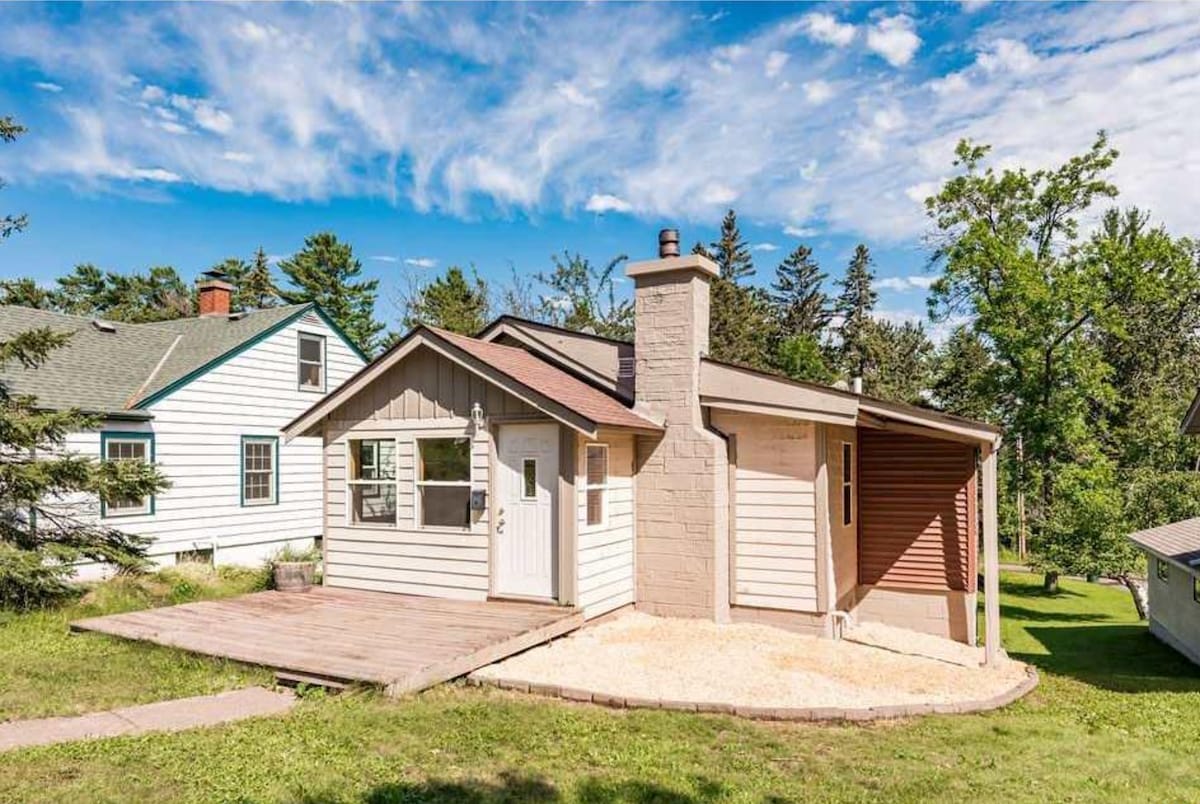A newly renovated cottage is presented with a light-colored exterior and a welcoming front porch. The structure features a chimney, and surrounding greenery enhances the tranquil setting. An expansive lawn provides a natural buffer between the cottage and neighboring homes.