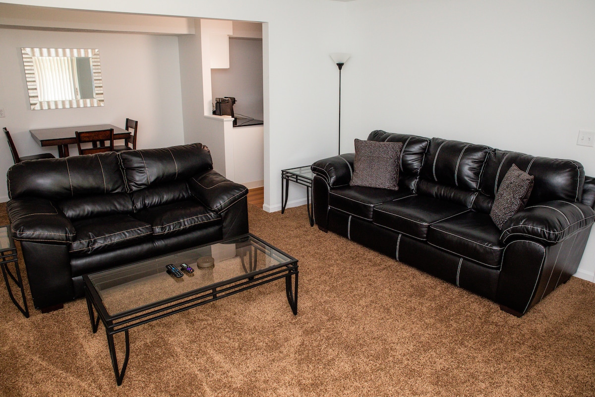 A living space features two black leather sofas positioned around a glass coffee table. The room is carpeted in a neutral tone, while a dining area with a table can be seen in the background. Natural light enters through a window with vertical blinds.