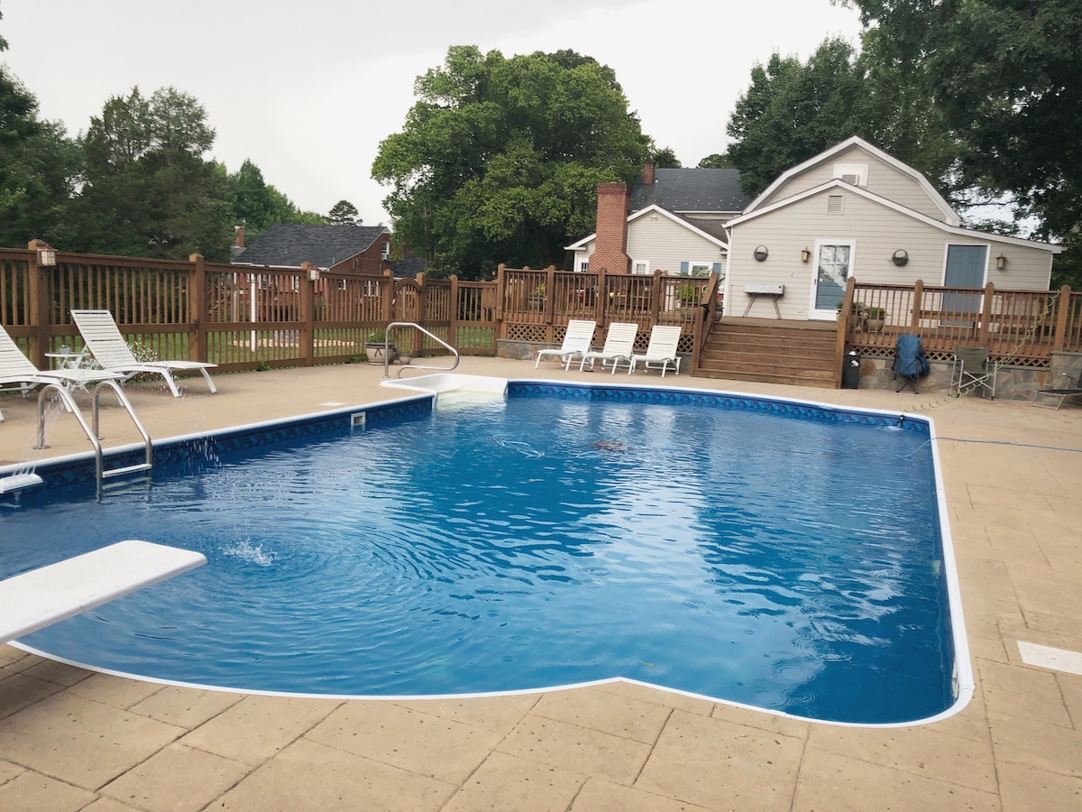 A large outdoor swimming pool with a diving board is surrounded by a spacious deck. Several lounge chairs are neatly arranged around the pool area. A two-story cottage is visible in the background, set against a backdrop of greenery.