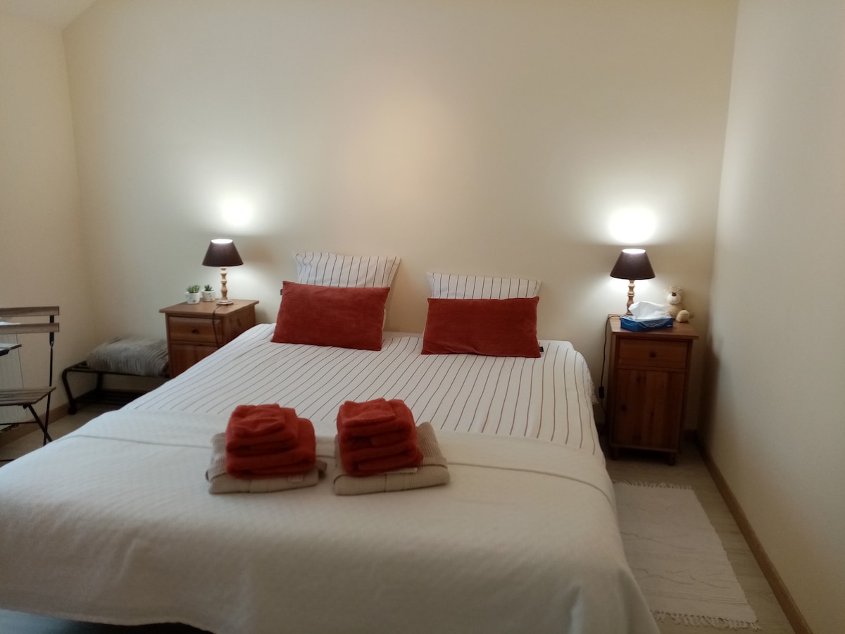 A spacious bedroom is featured with a large bed topped with white linens and two orange accent pillows. Nightstands with lamps are placed on either side of the bed, and neatly folded towels are arranged at the foot of the bed.
