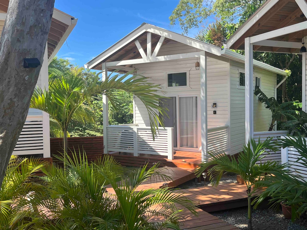A private casita is surrounded by lush tropical plants and wooden decking. Natural light filters through large windows, creating an airy ambiance. The structure features a welcoming porch with decorative railings, emphasizing the connection between indoor and outdoor living.