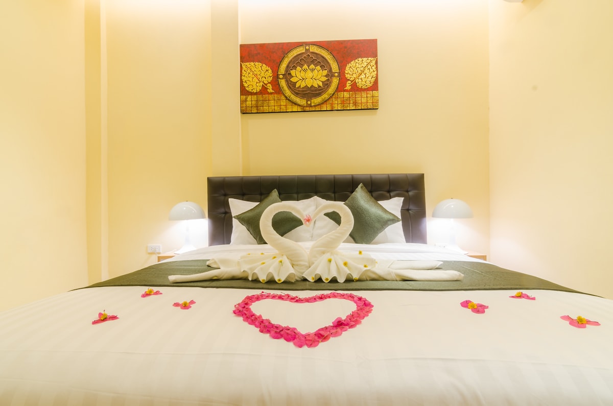 The room features a double bed adorned with a decorative arrangement of towels shaped like swans. A heart-shaped pattern made of flower petals is displayed on the bedding. Warm light is provided by two bedside lamps, while artwork adds color to the wall above the bed.