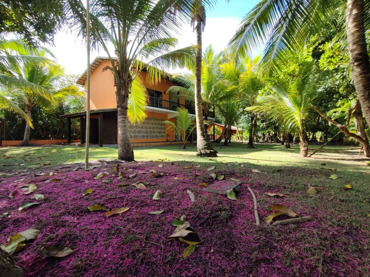 A spacious area is surrounded by palm trees, with a gently sloping garden covered in vibrant purple foliage. The orange building stands in the background, featuring a large balcony and multiple openings that blend with the natural environment.