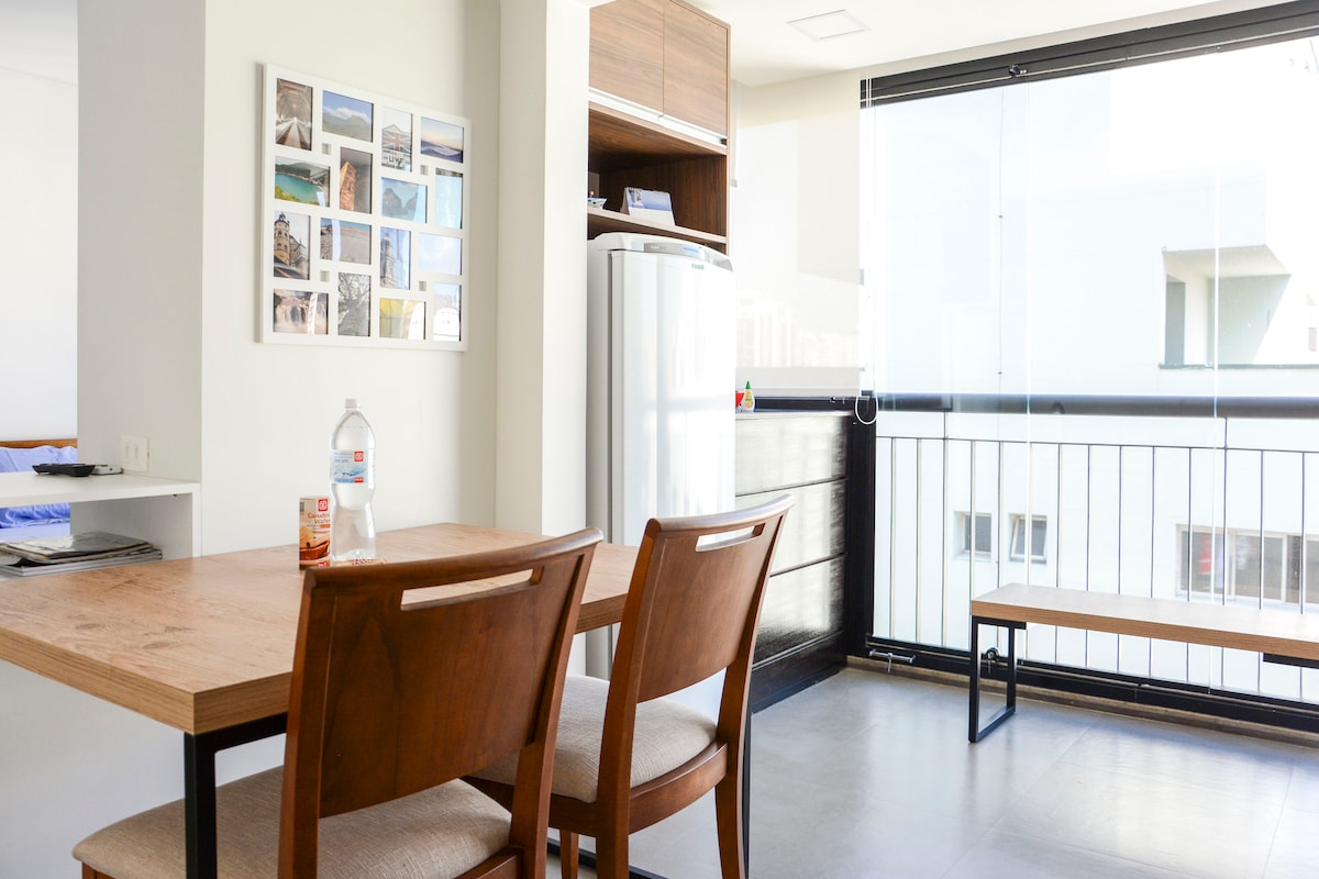 A modern dining area features a wooden table with two chairs, accompanied by a bottle of water. A gallery wall displays a collection of framed photos. In the background, a sleek refrigerator is partly visible, alongside a minimalist bench by a glass door leading to an outdoor space.