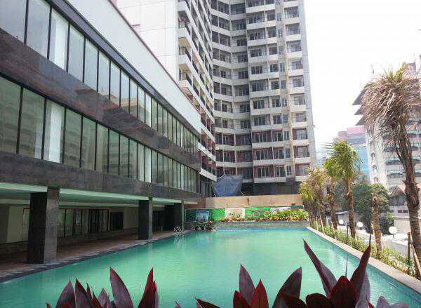 A large swimming pool is beautifully set against a backdrop of modern apartment buildings. Lush greenery and palm trees frame the pool area, which is highlighted by clear blue water reflecting the surrounding architecture.