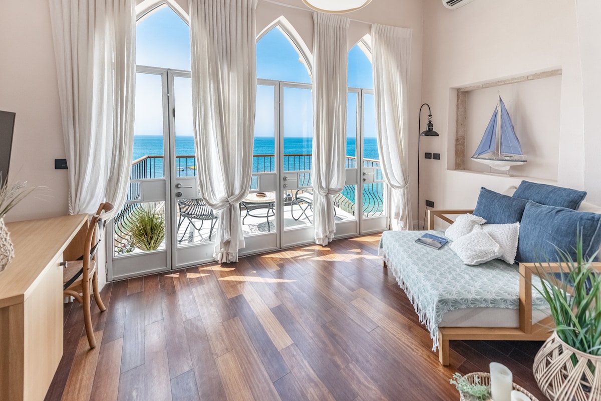A bright living area features large arched windows offering sweeping views of the sea. A comfortable sofa is paired with a light-colored throw and decorative pillows. The wooden floors create a warm contrast to the soft color palette, while a small dining set is visible on the balcony.