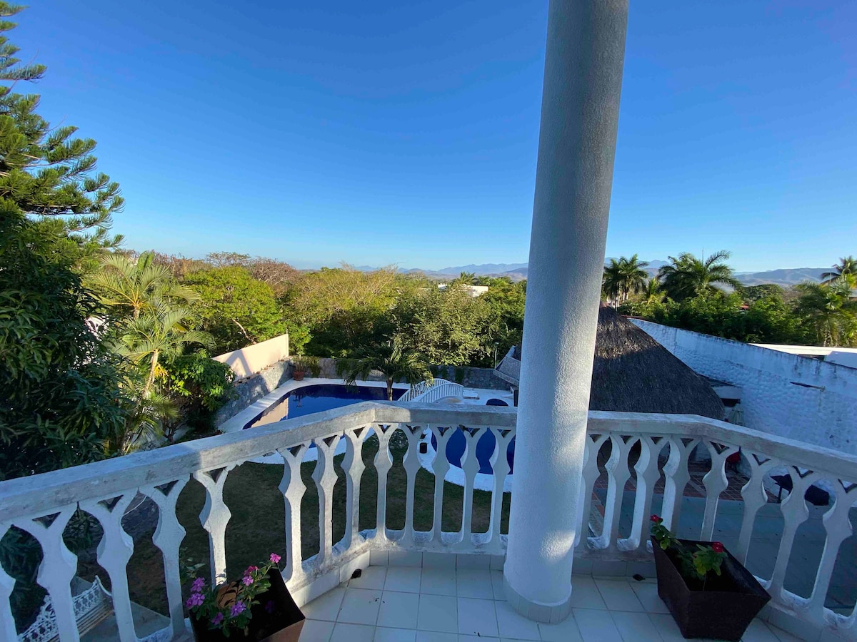 A balcony view is provided, showcasing a garden surrounded by trees and a clear blue sky. A pool with a thatched-roof structure is visible below, offering a serene outdoor space for relaxation.