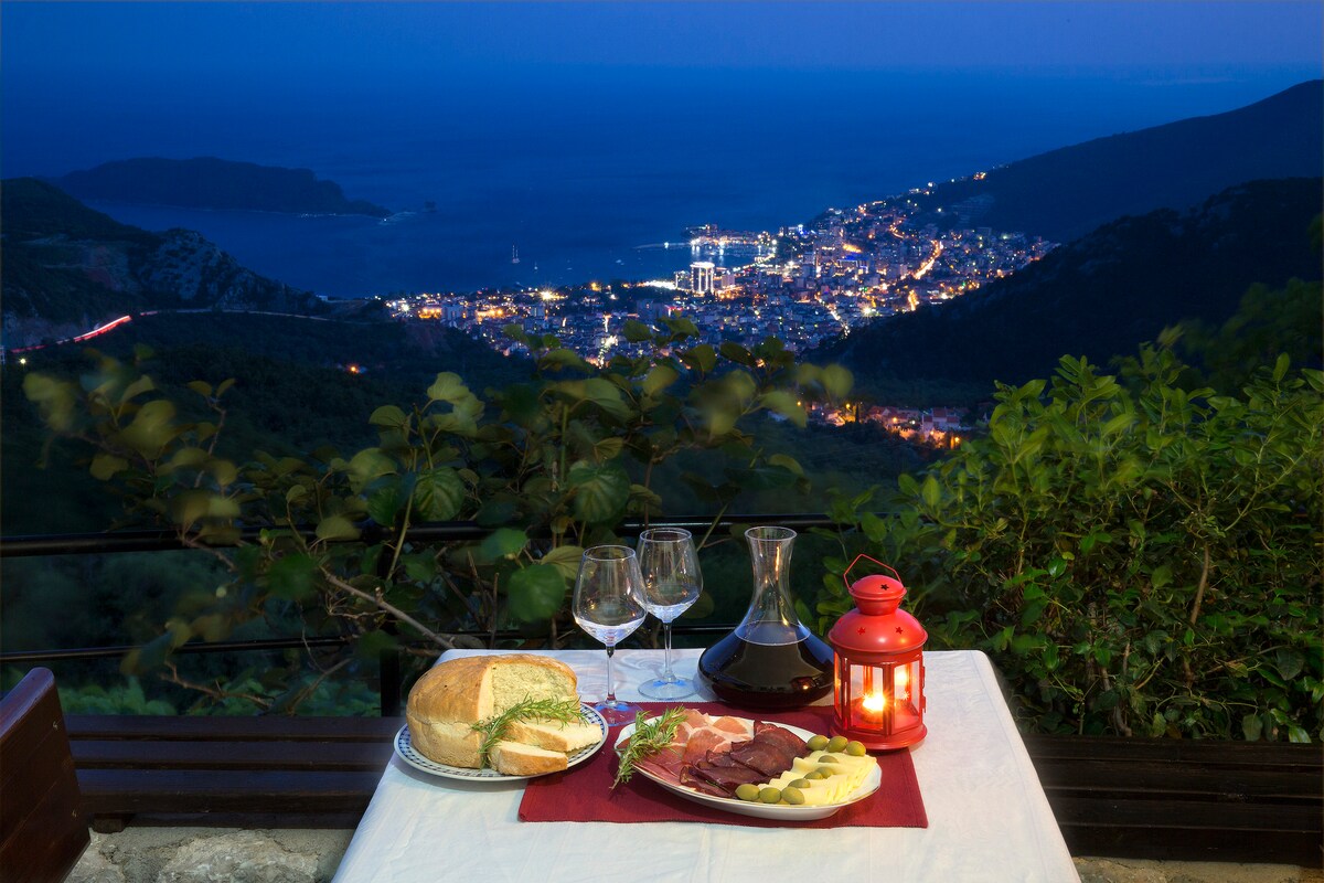 A table is set with a white tablecloth, featuring a selection of bread, charcuterie, and cheese, alongside two wine glasses and a carafe. A red lantern provides soft lighting, with a panoramic view of the illuminated Budva cityscape and the sea in the background.