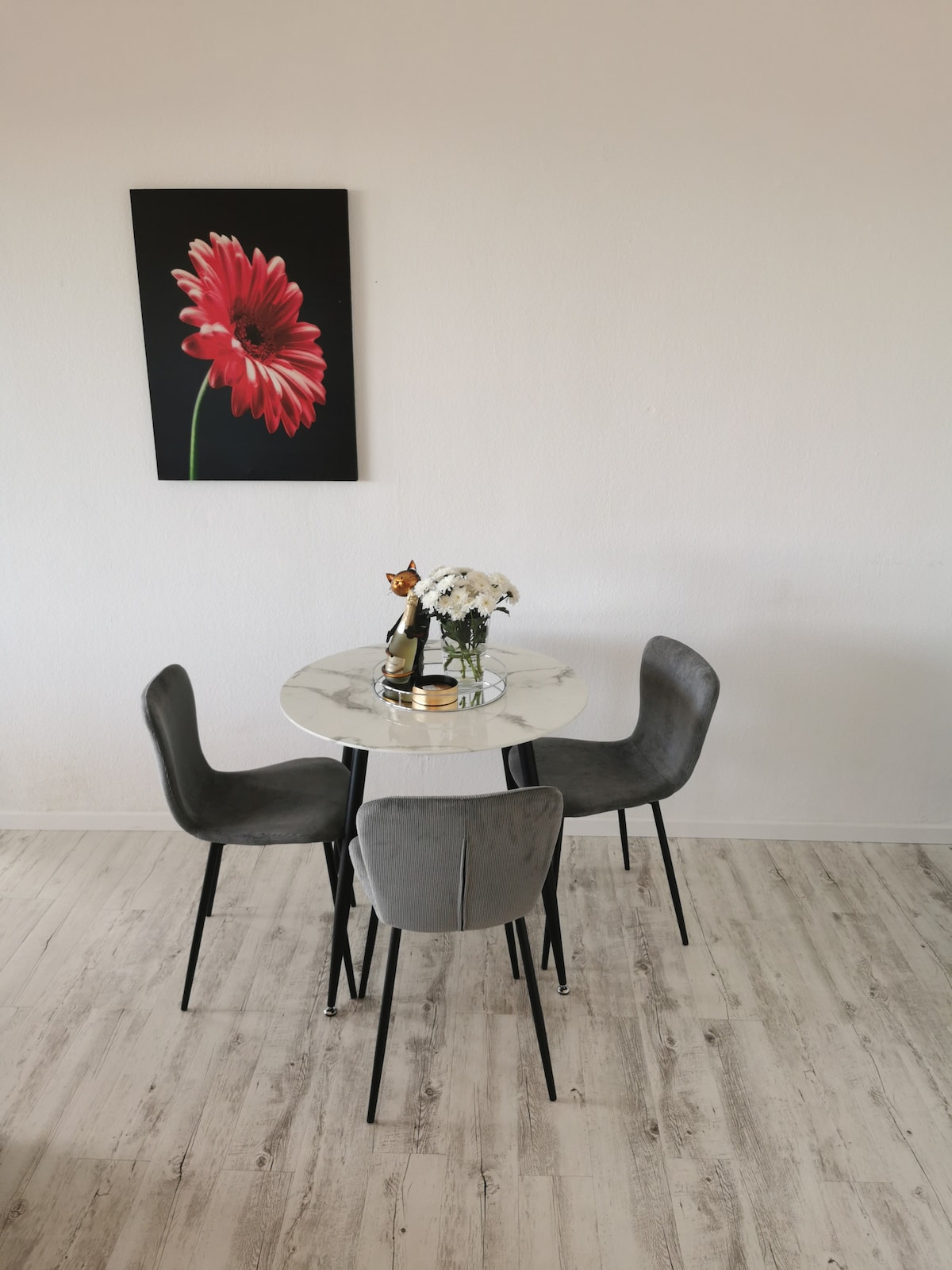 A round marble dining table is surrounded by four grey upholstered chairs. A small vase of white flowers is placed on the table, adding a touch of greenery. A vibrant red flower painting is displayed on the wall, contributing to the space's design.