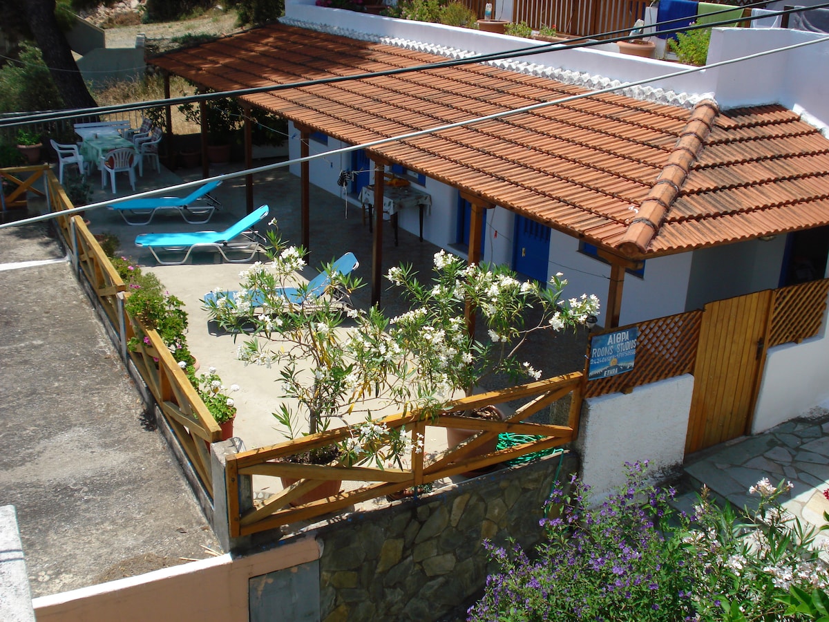 A spacious outdoor area is presented, featuring sunbeds and a wooden fence. The shaded veranda, outfitted with a tiled roof, offers a seating area. Surrounding greenery enhances the environment, with a small garden area visible. The entrance to the apartment is accessible from this space.