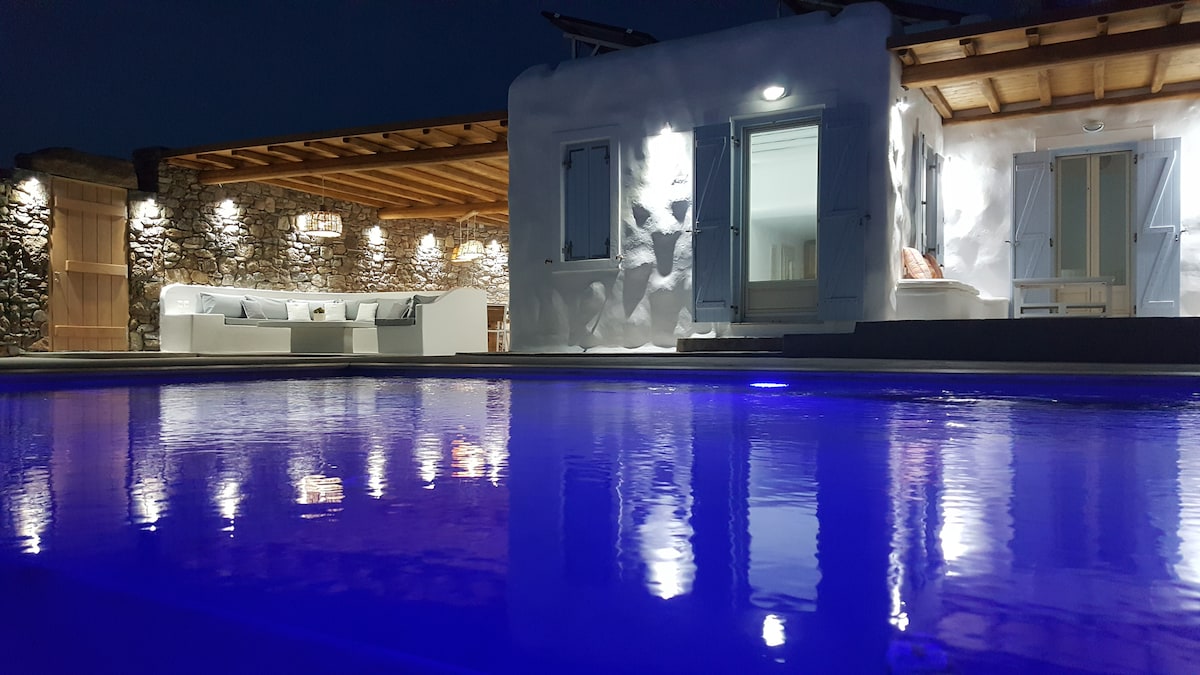 An outdoor area is illuminated by soft lights, reflecting off a tranquil pool. The villa features a stone facade and glass doors, with a comfortable seating area visible under a wooden pergola. The serene atmosphere is enhanced by the night sky.