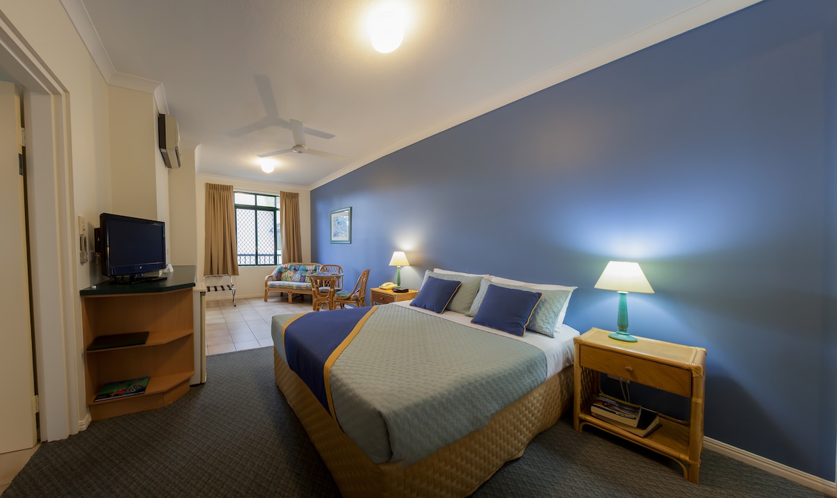 A spacious bedroom features a large bed with soft bedding and two bedside tables, each with a lamp. A small seating area with a rattan chair is set against a blue accent wall, while a television and dresser are positioned nearby.