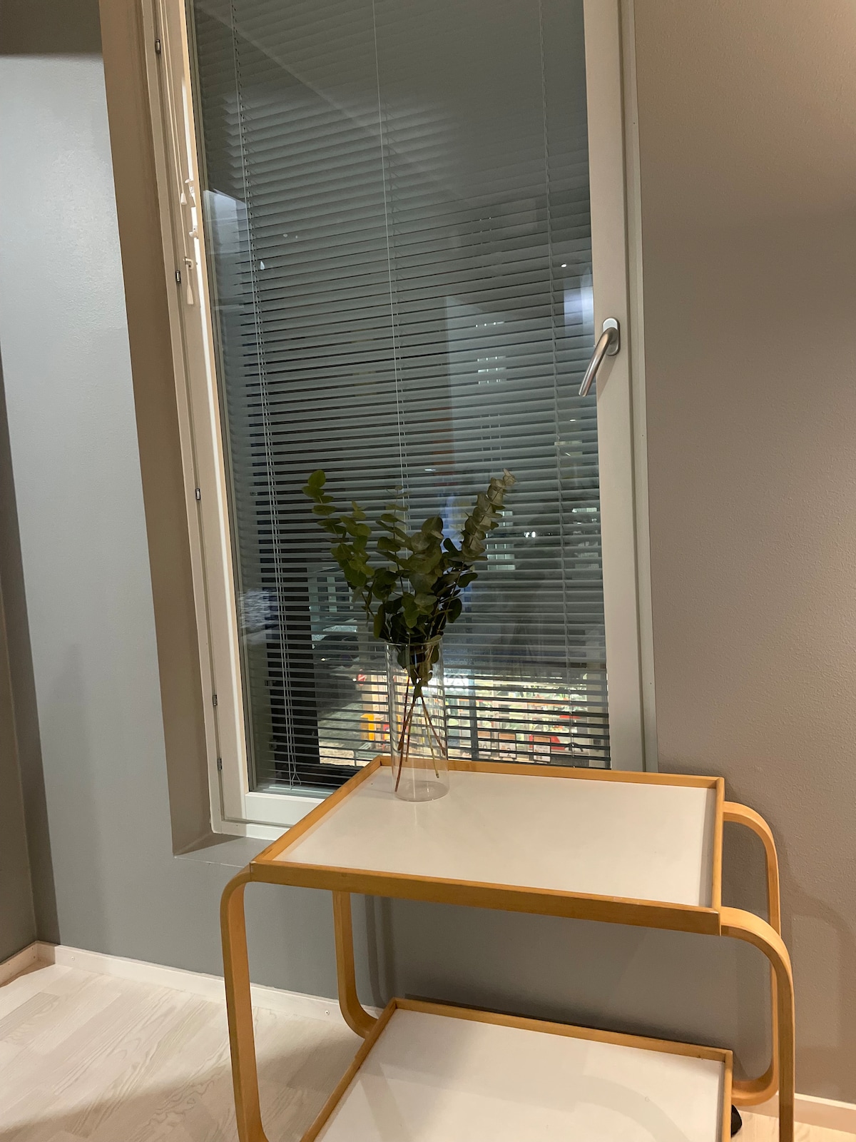 A small table with a light wooden frame is positioned beside a window. A clear vase containing greenery rests atop the table. The window features horizontal blinds, allowing soft light to filter through while providing a view of the city through the glass.