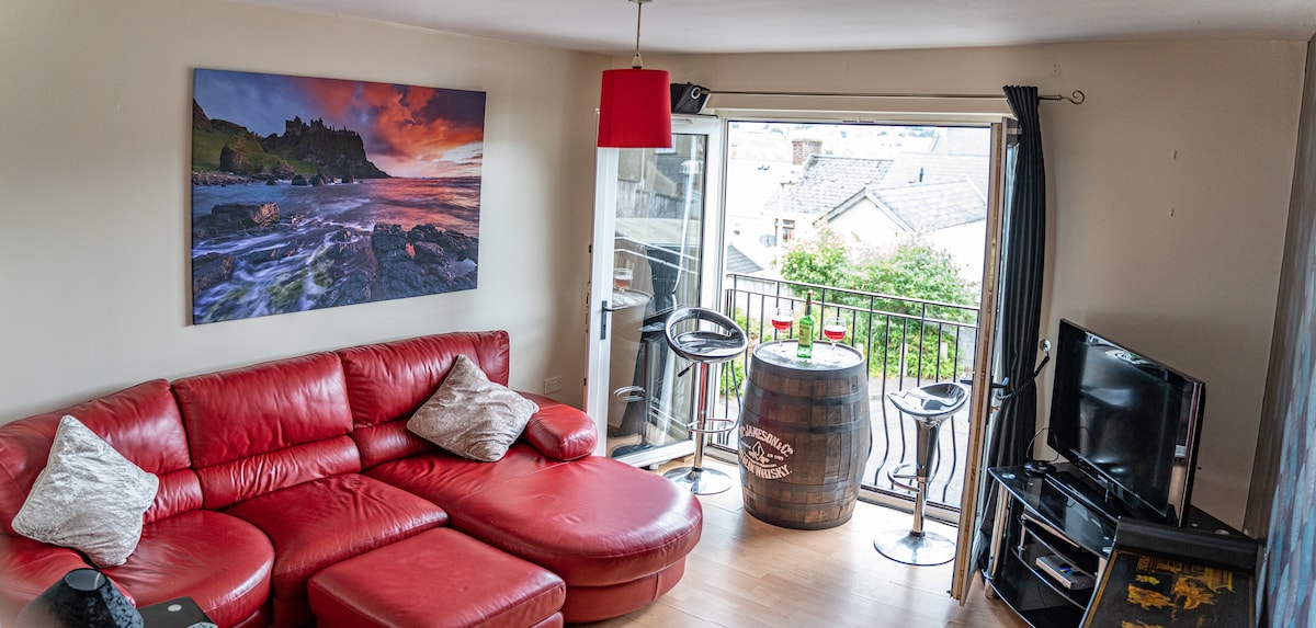 A spacious living area features a red sectional sofa accompanied by two decorative pillows. A vibrant landscape photograph is displayed on the wall. Patio doors lead to a small balcony with two barstools and a table, providing a view of greenery outside.