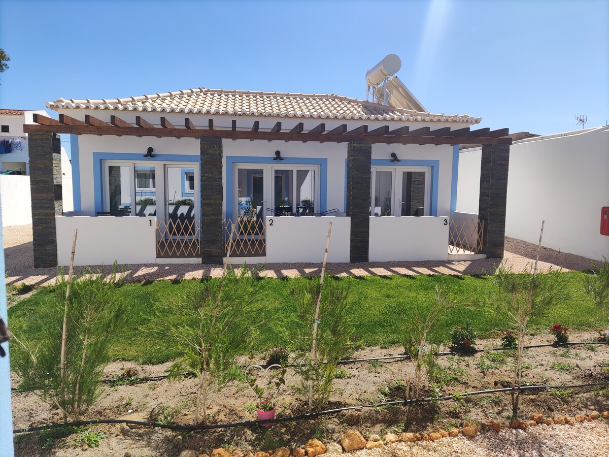 The exterior of a modern accommodation showcases three independent entrances, framed by a shaded veranda. A well-maintained garden with green grass and small plants lies in front, complementing the light-colored walls and stone accents of the property.