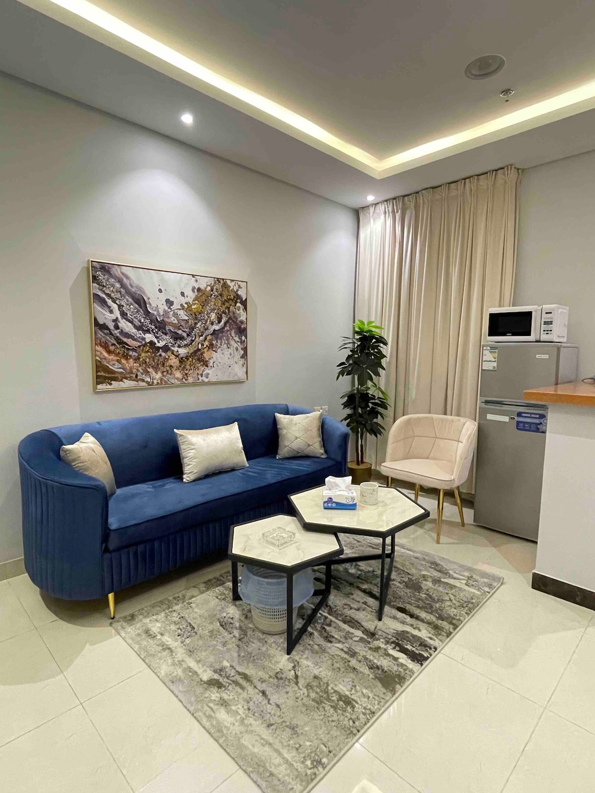 A cozy living space features a blue velvet sofa with decorative pillows, accompanied by a light-colored armchair. A small coffee table sits on a textured rug, while a modern painting adorns the wall. A kitchenette with a refrigerator and microwave is visible in the background.