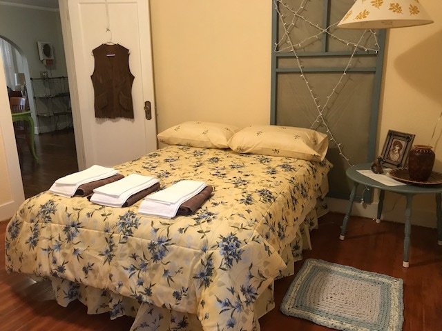 A cozy bedroom features a floral-patterned bedspread over a spacious bed with two neatly arranged pillows. Fresh towels are placed at the foot of the bed. A lamp and a small side table with a framed photo enhance the charm of the room, complemented by warm wooden flooring.