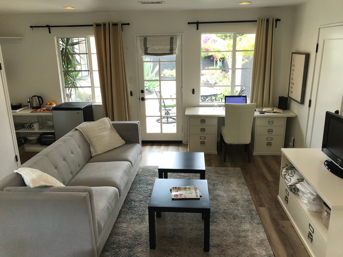 A cozy guest studio features a gray sofa and two low tables on a light area rug. Natural light fills the room through large windows adorned with beige curtains. A desk with a computer overlooks a shared courtyard, visible through glass doors.