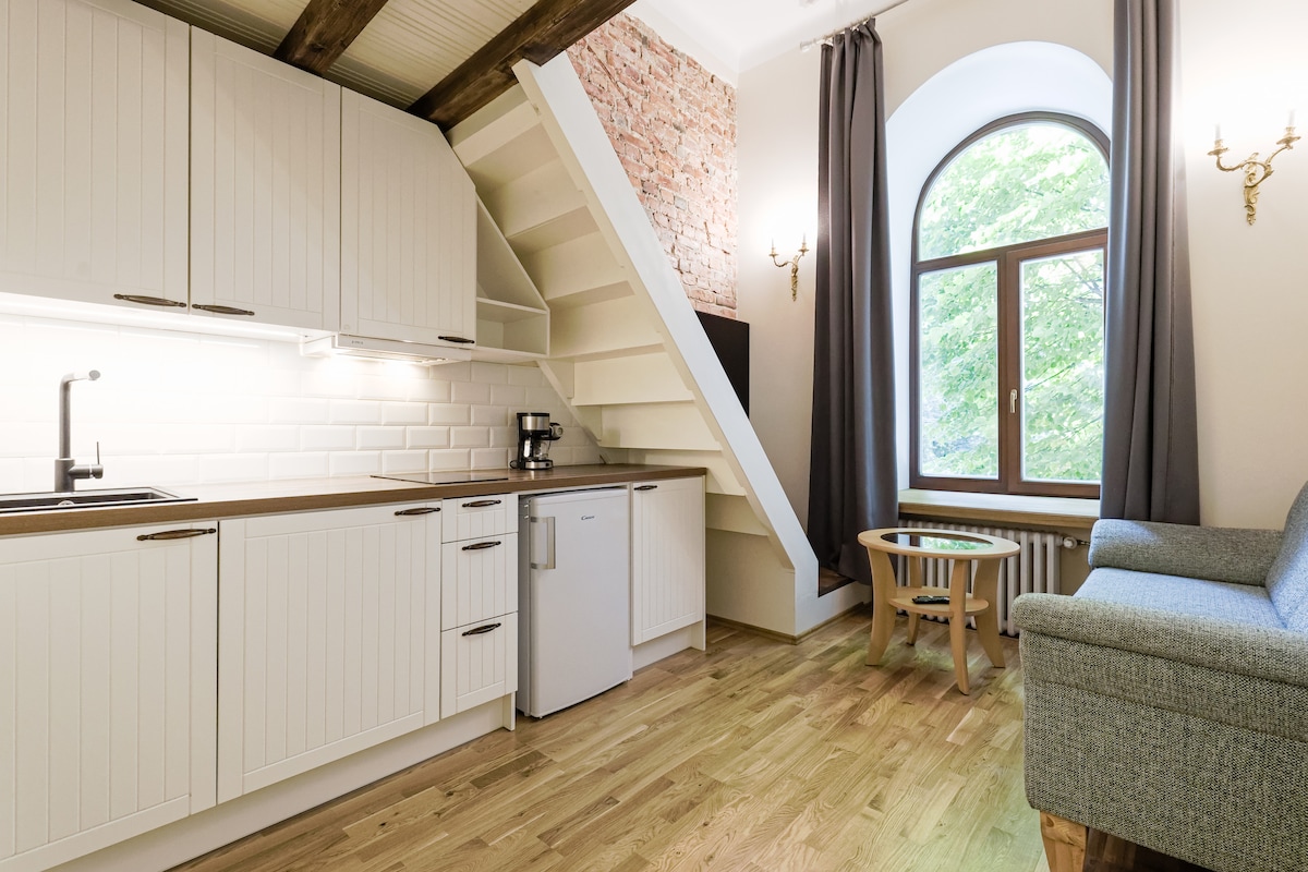The kitchen area is equipped with modern amenities, including a refrigerator and coffee maker, set against a backdrop of exposed brick walls. A comfortable sofa is positioned near a large window, allowing natural light to fill the space. Wooden flooring enhances the inviting ambiance.