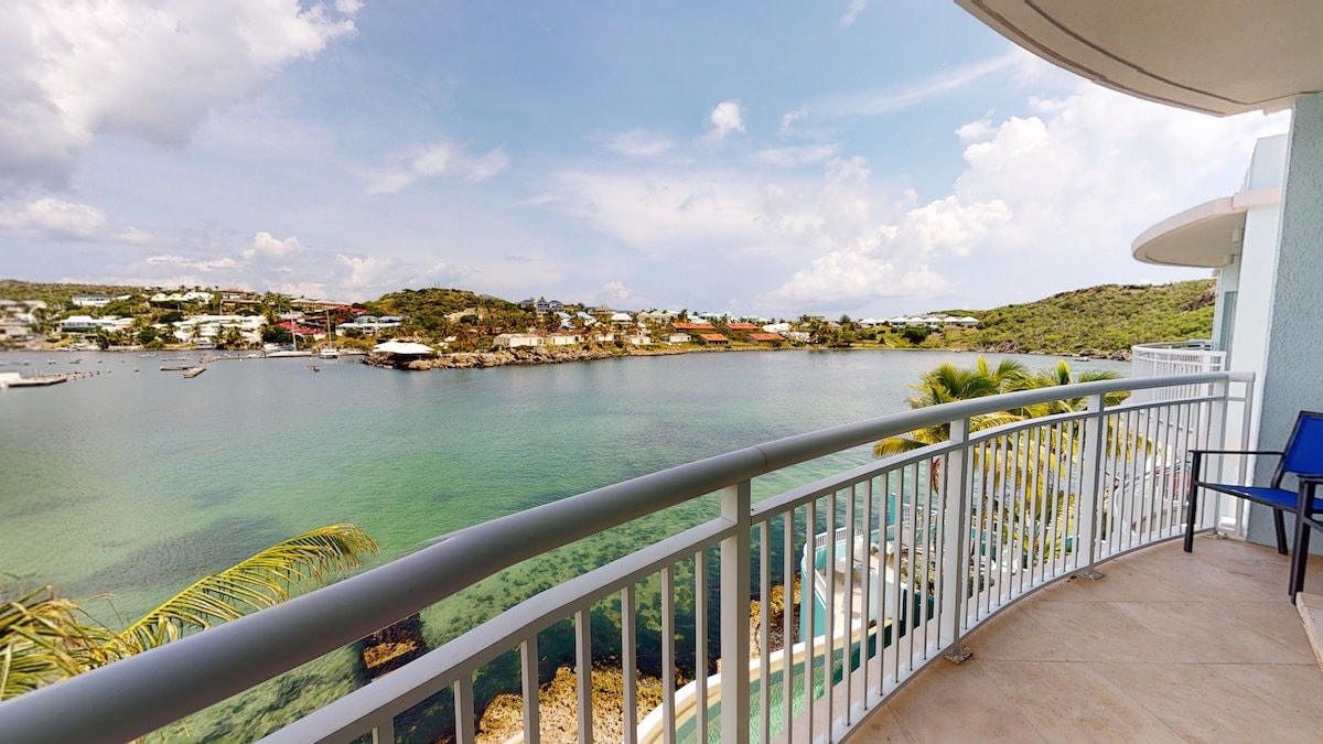 A wide balcony offers views of the marina and lagoon, with calm waters reflecting the sky. Surrounding green hills and coastal buildings create a scenic backdrop. Outdoor seating is provided, enhancing the space for relaxation.