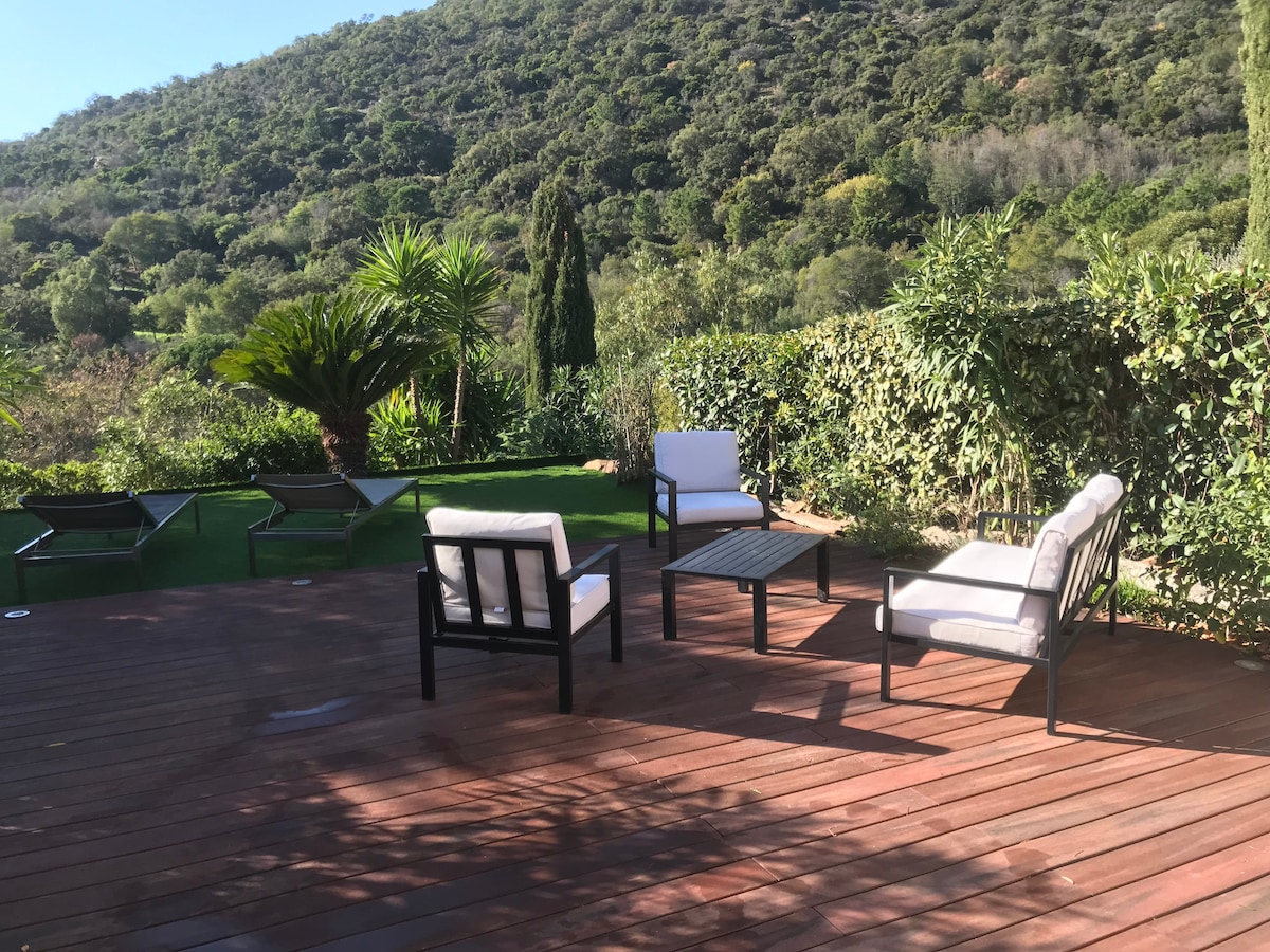 A spacious wooden deck is furnished with four modern seating arrangements, consisting of two armchairs and a coffee table. Lush greenery and palm trees surround the area, creating a serene natural backdrop. In the distance, lounge chairs are visible on the grass.