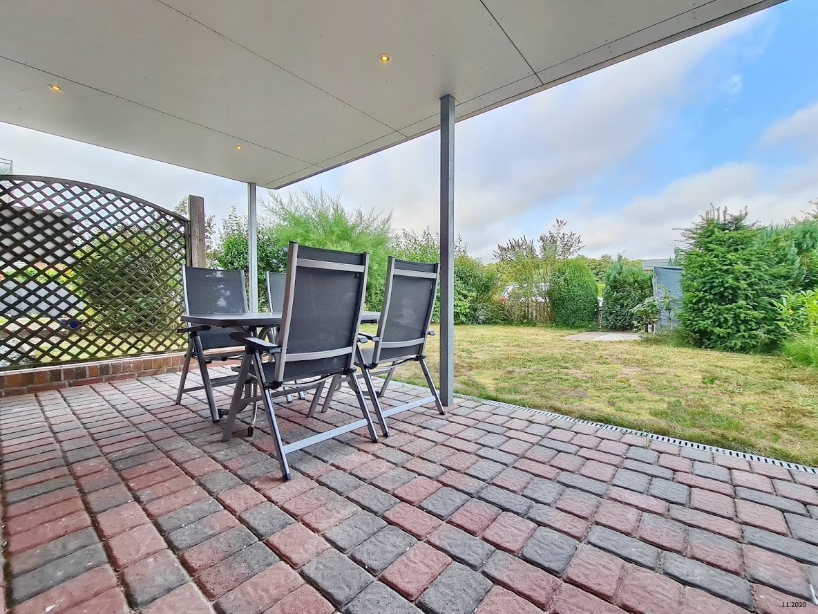 A covered outdoor terrace is featured, furnished with a set of four black chairs arranged around a small table. The space is surrounded by greenery and garden beds, with a stone pathway leading to the grassy area beyond.