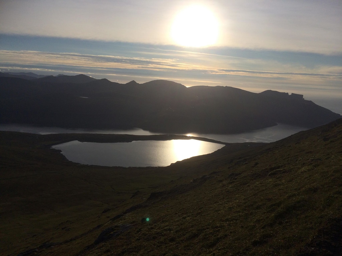 A serene landscape captures rolling hills leading down to a calm lake, reflecting sunlight. Mountains rise in the background against a clear sky, creating a peaceful natural scene. The sun casts a gentle glow over the water, enhancing the tranquil atmosphere.