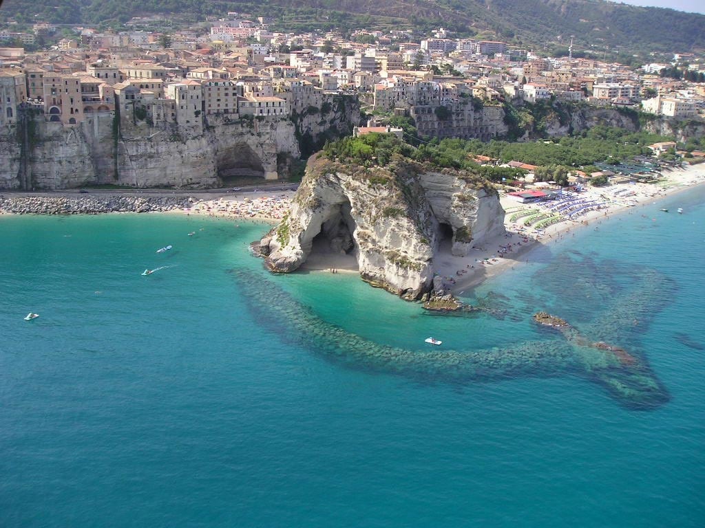 This aerial view showcases a scenic coastline featuring clear turquoise waters and expansive sandy beaches. A rocky promontory with lush greenery juts into the sea, while beachgoers are seen enjoying the sun. The backdrop consists of charming coastal buildings and lush hills.
