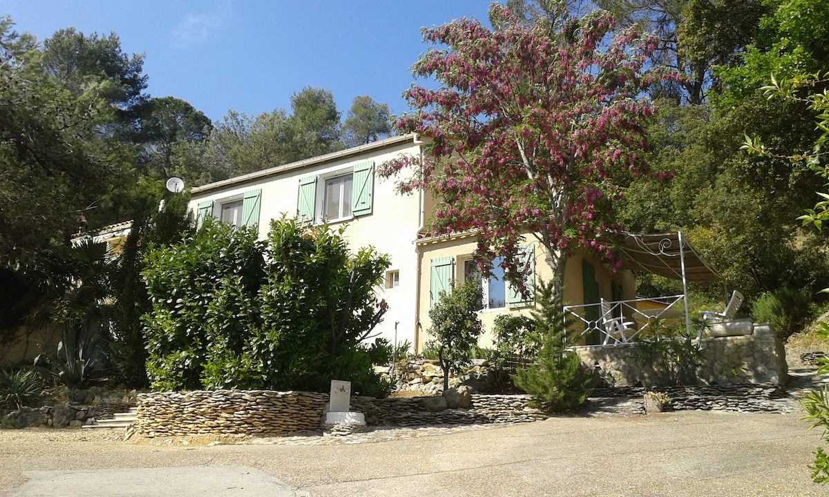 A charming two-story house is nestled among trees and shrubs, showcasing light-colored walls and green shutters. A covered terrace with outdoor furniture is visible, surrounded by a landscaped garden. The driveway leads to the entrance, enhancing the tranquil setting.