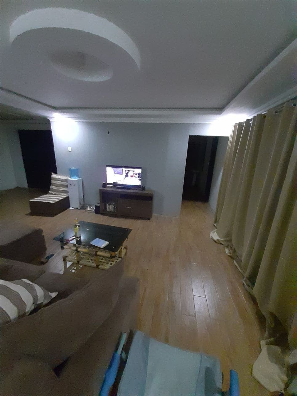 A spacious living area features a television positioned on a low stand. A comfortable sofa, partially visible, includes striped cushions. Natural light enters through a window with light brown curtains, and a beverage cooler rests against a wall. A wooden floor adds warmth to the setting.