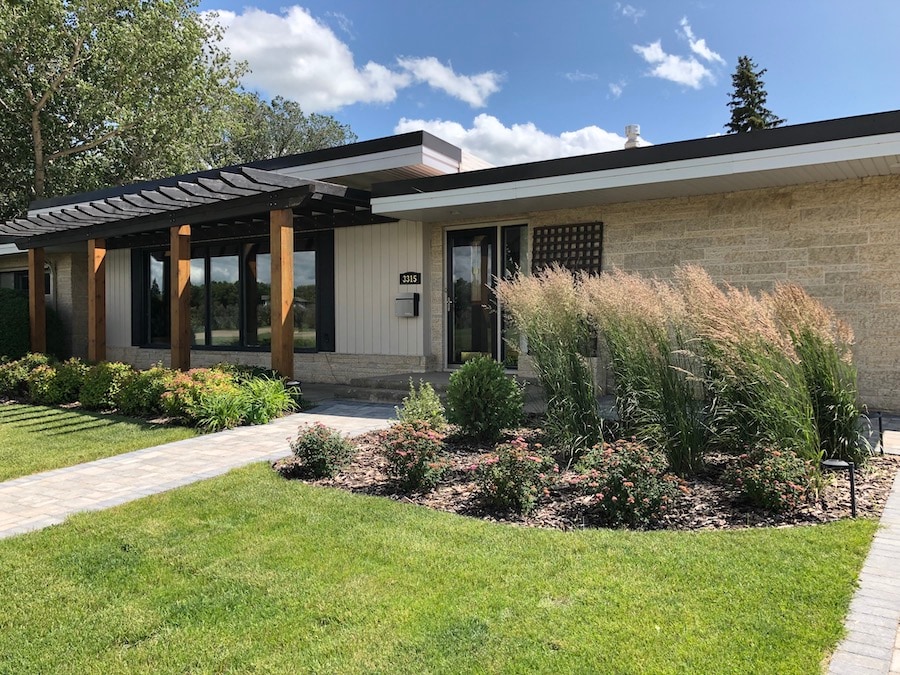 The front exterior of the mid-century modern bungalow showcases a landscaped yard with a variety of plants and flowers. Large windows allow natural light to enter, while a pathway leads to the entrance. The building features a blend of stone and wood elements under a blue sky.