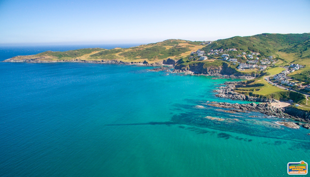 The image captures a sweeping aerial view of the coastline, featuring a blend of rocky outcrops and gentle waves. The deep blue water contrasts with the lush green hills in the background, showcasing a serene and expansive natural landscape.