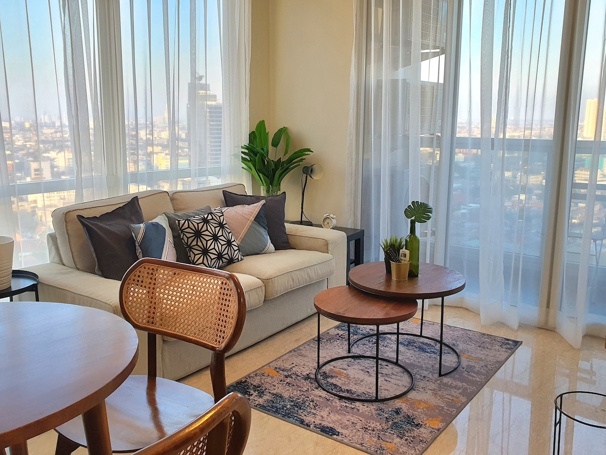 A modern living area is featured with a comfortable sofa adorned with cushions. Two round coffee tables are positioned on a soft area rug, while large windows draped with sheer curtains allow natural light to fill the space, revealing a view of the city skyline.