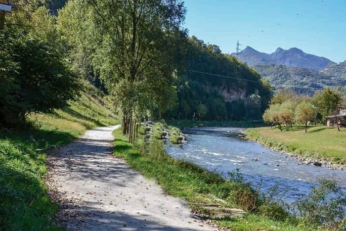 A serene riverside path meanders alongside a gently flowing river, bordered by lush greenery and trees. In the background, mountains rise majestically under a clear blue sky, completing the natural scenery.