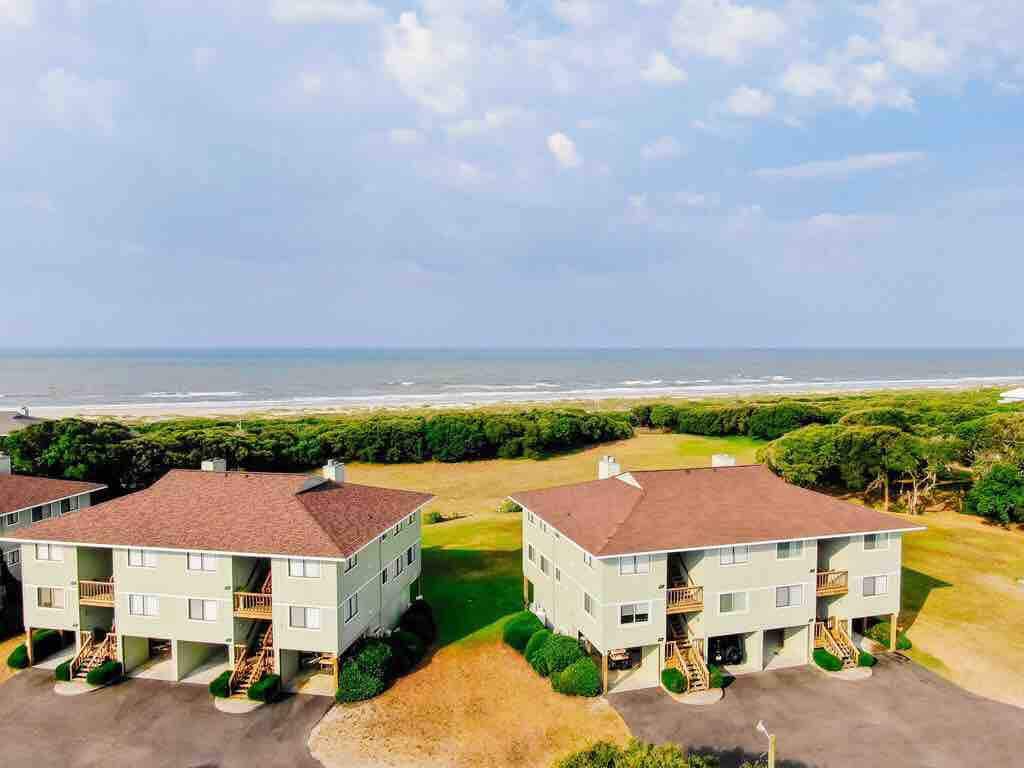 Two two-story buildings are visible, situated amidst well-maintained green landscaping. The ocean is seen in the background, with gentle waves lapping at the shore. A vast open space in the foreground enhances the serene coastal setting.