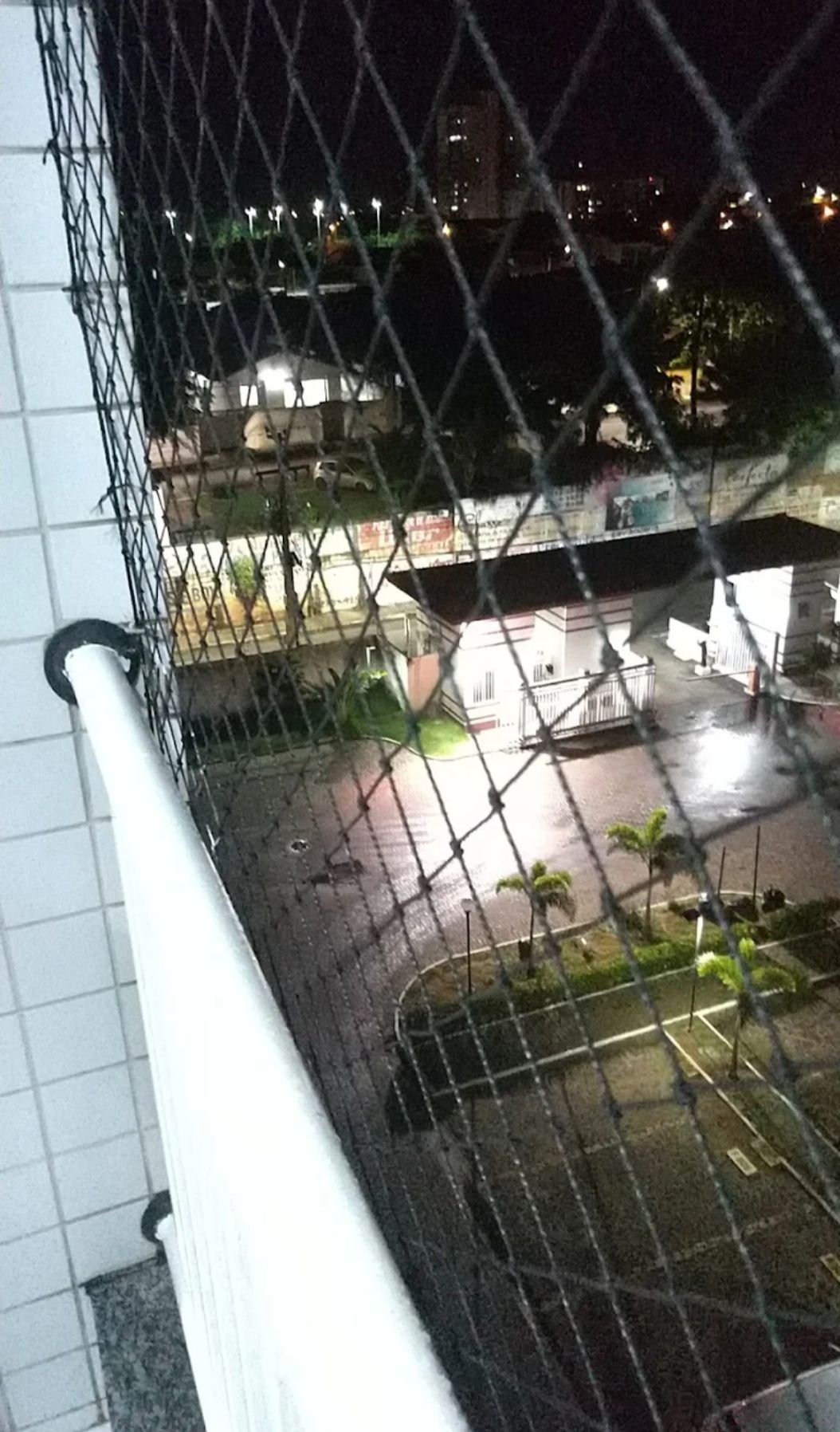 A nighttime view from a balcony showcases a well-lit courtyard below, featuring pathways and green plants. A protective netting frames the image, emphasizing the height of the building. Nearby illuminated spaces suggest a vibrant atmosphere, with urban structures visible in the background.